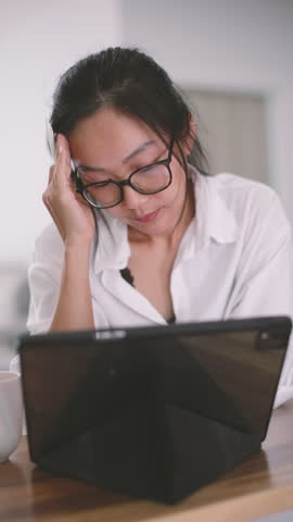 Tired Asian woman holds pen and touches forehead while looking at tablet. Creative burnout moment in vertical modern workspace, wearing eyeglasses and casual white shirt with soft light.