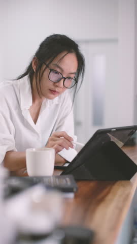 Asian woman in eyeglasses leans on hand, deeply focused on tablet during remote work. Casual white shirt, coffee cup, and vertical home office setup create calm, creative workspace mood.