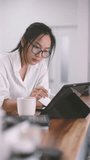 Asian woman in eyeglasses leans on hand, deeply focused on tablet during remote work. Casual white shirt, coffee cup, and vertical home office setup create calm, creative workspace mood. - Powered by Shutterstock - Get 15% off with code: PIKWIZARD15