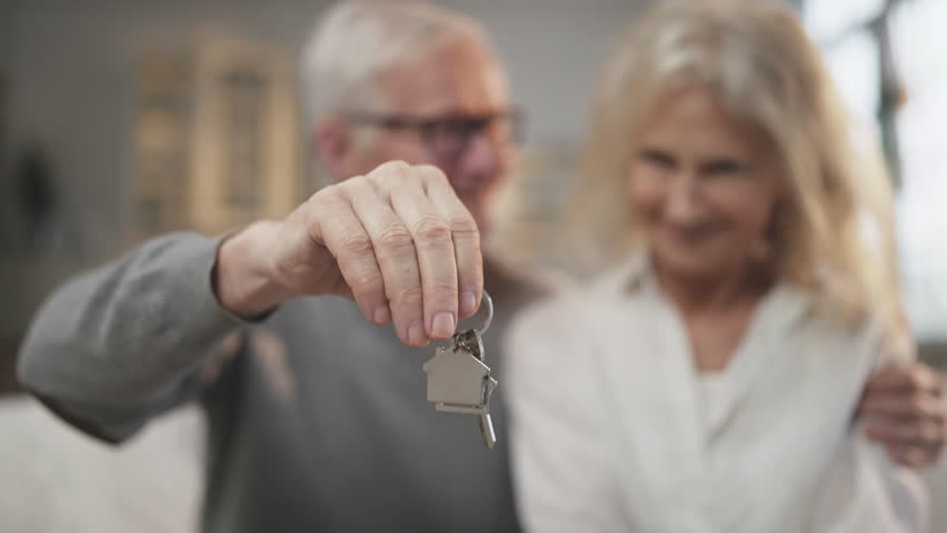 Buy new apartment, keys in hands of elderly man, blurred portrait of aged couple . Mature spouses rejoicing by finish of mortgage, happy house owners, real estate agency, rent, purchase or sale flat