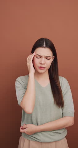 Vertical clip. Young woman with migraine headache. Isolated on brown background in studio