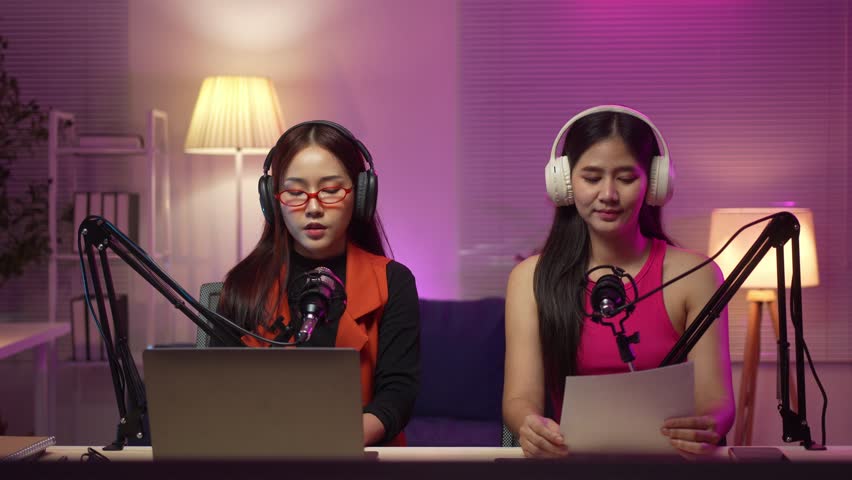 Two women are recording a podcast in a room with a laptop and a desk lamp. They are wearing headphones and microphones