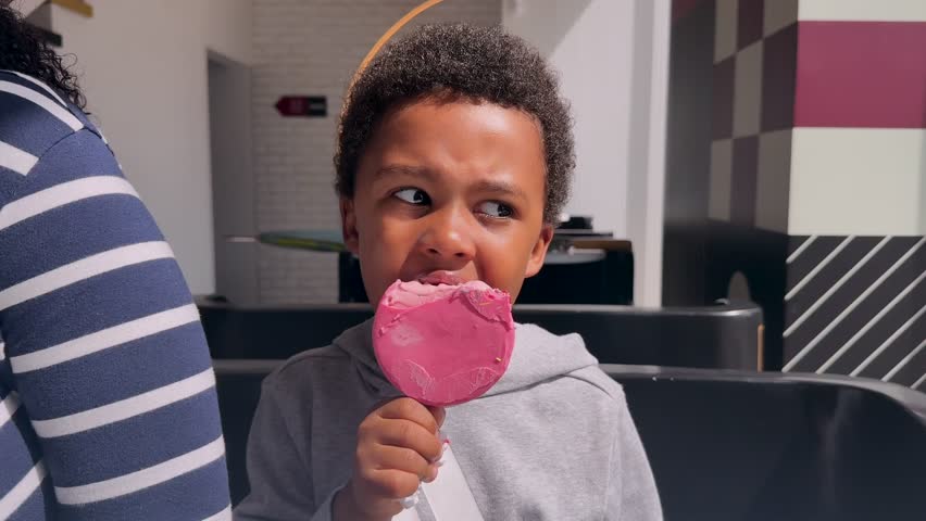 Close-up view of a young biracial black boy eating a pink strawberry-flavored artisan ice cream indoor next to his mother, wearing a gray hoodie