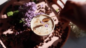 A woman stirs a steaming cup of coffee with milk and frothy foam using a spoon, placed on a wicker tray next to lilac flowers in a brown craft envelope. A serene mix of floral elegance and aromatic - Powered by Shutterstock - Get 15% off with code: PIKWIZARD15