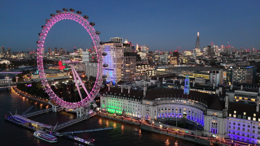  Stunning aerial footage of the London Eye glowing in pink during a vibrant blue sunset. The iconic Ferris wheel stands out against the backdrop of the London skyline, with the River Thames and city