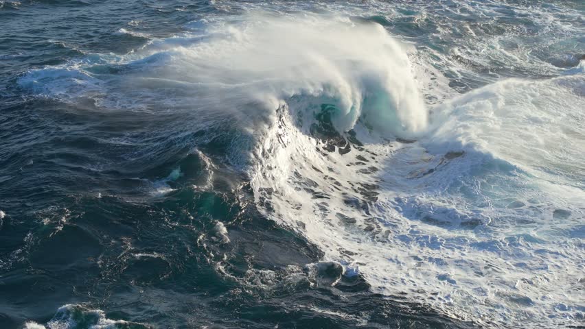 Massive ocean waves crash against rocky shore, creating impressive splashes and mist near coastal cliffs. Aerial slow motion shot of big epic waves surf crash