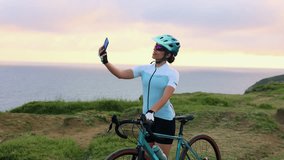Young Latin woman cyclist wearing helmet and technical clothing takes a selfie with her smartphone standing next to her gravel bike by the sea at sunset - Powered by Shutterstock - Get 15% off with code: PIKWIZARD15