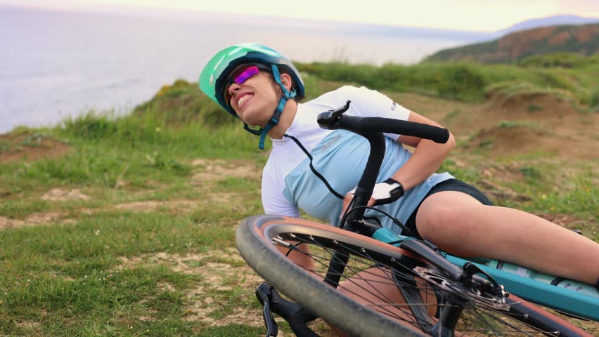 Latin cyclist woman wearing helmet and sunglasses falling from her gravel bike on a grassy cliff near the sea at sunrise