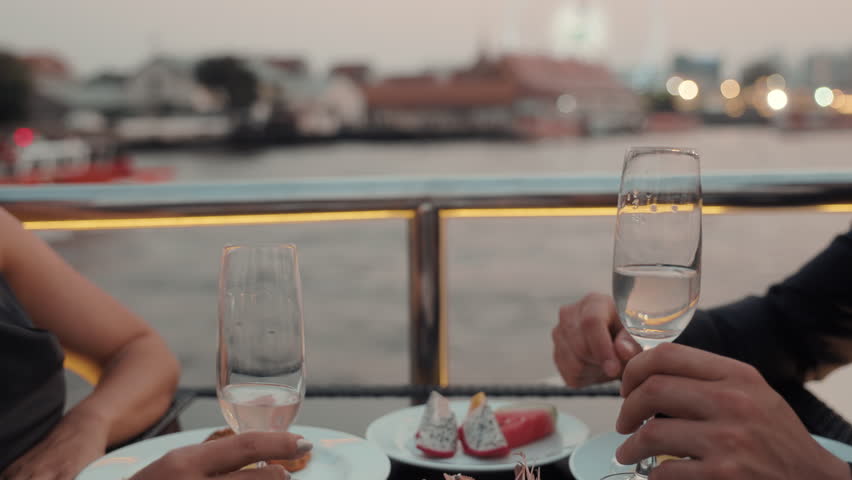 Romantic partners hands clink glasses of expensive wine on dinner riding luxury yacht closeup. Young couple celebrates anniversary sailing modern ship on sea journey in evening