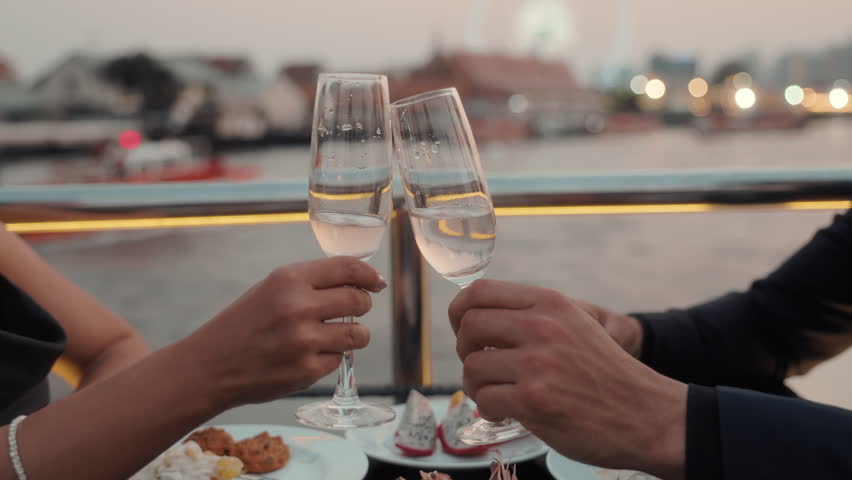 Romantic partners hands clink glasses of expensive wine on dinner riding luxury yacht closeup. Young couple celebrates anniversary sailing modern ship on sea journey in evening