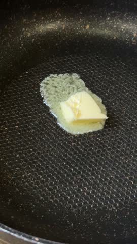 A piece of butter slowly melting on a hot nonstick frying pan. The video shows the start of cooking, with the butter beginning to sizzle and spread on the pan’s surface.