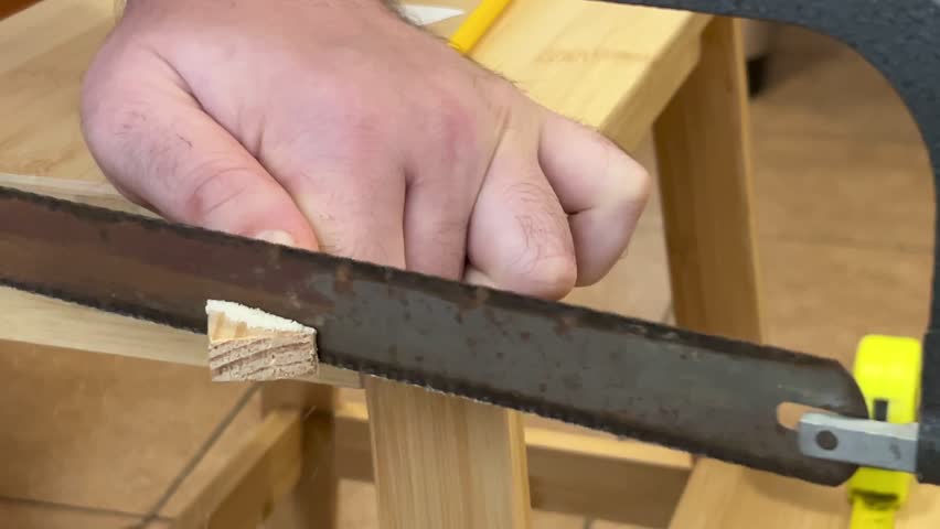 Detail view of woodworking process. A person uses a handsaw to cut a small wooden block on a home workshop bench.