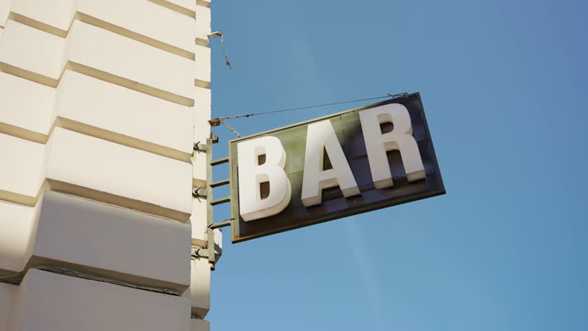 Sign of a local bar in small town or city with blue sky outside. Drinking establishment place with big street sign in corner in main street or downtown during afternoon or weekend. Low angle view
