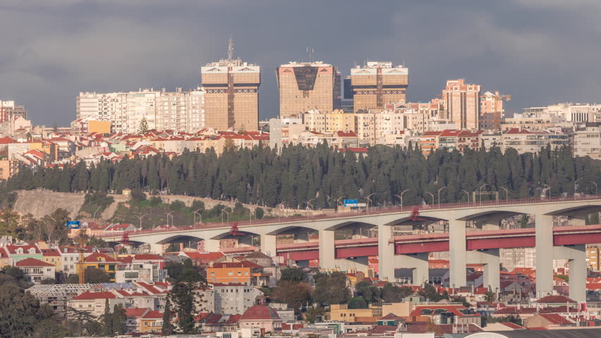 Aerial timelapse of Lisbon skyline featuring colorful houses, Amoreiras shopping center towers and the 25th of April Bridge. Historic district with green trees and Prazeres Cemetery in the foreground