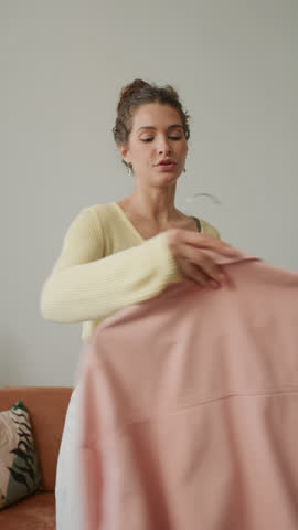 Young beautiful girl presenting light pink shirt and flower patterned dress, sharing outfit ideas during fashion vlog recording at home. Vertical format clip