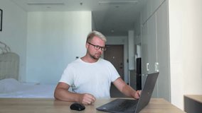 A young guy , blond, in a white t-shirt sits at a desk in a stylish flat and works on his laptop. The laptop breaks down, the man doesn't know what to do and gets angry - Powered by Shutterstock - Get 15% off with code: PIKWIZARD15