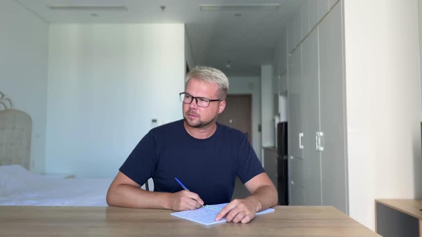 A young guy, dyed stylish blond in a blue t-shirt sits at a desk in a bright interior and writes in a notebook. He doesn