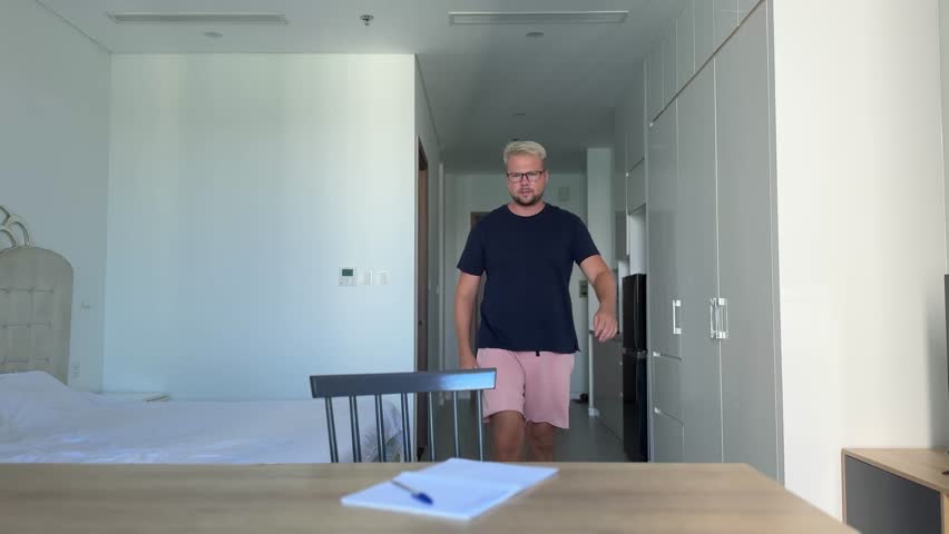 A young guy, dyed stylish blond in a blue t-shirt sits at a desk in a bright interior and writes in a notebook. He doesn