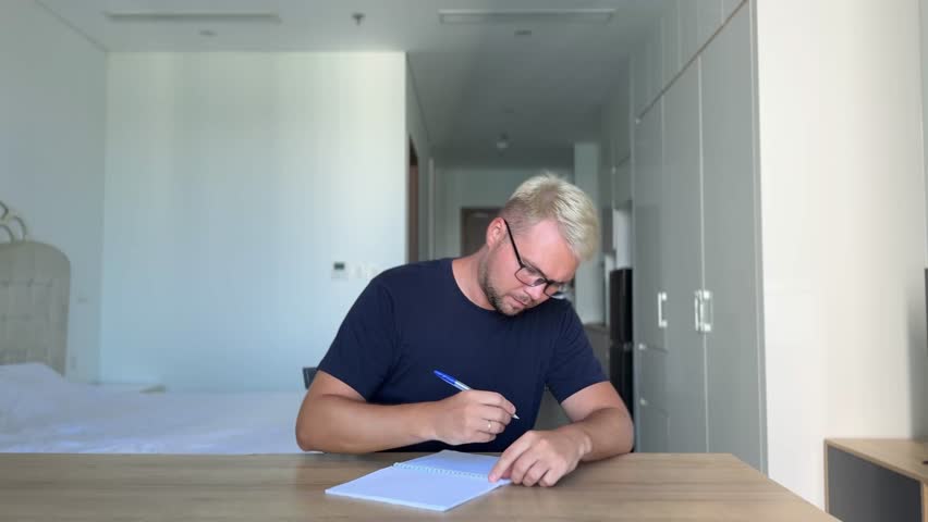 A young guy, dyed stylish blond in a blue t-shirt sits at a desk in a bright interior and writes in a notebook. He doesn