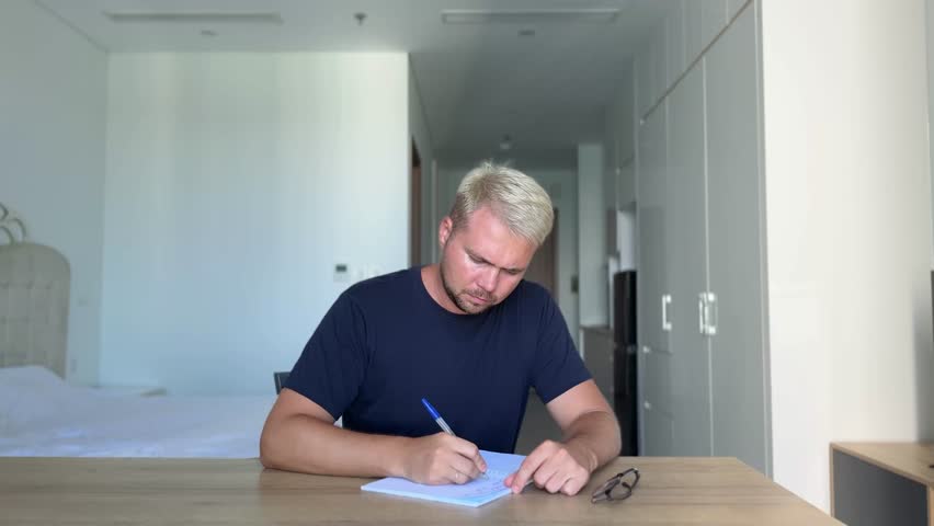 A young guy, dyed stylish blond in a blue t-shirt sits at a desk in a bright interior and writes in a notebook. He doesn