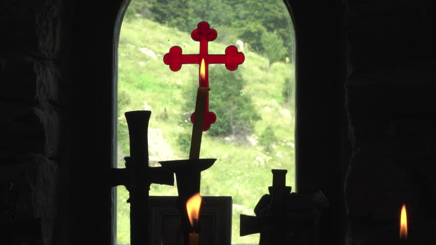 View from inside out through the window of a Bulgarian Orthodox Chapel. Burning candles against a background of a red cross painted on glass. Eastern Orthodox Chapel high in the mountains. Silhouette.