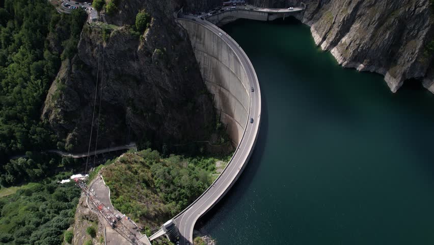 Drone shot of cars driving on Vidraru Dam in Romania, circling left. Blue lake, dramatic curves, and green mountain backdrop in 4K.