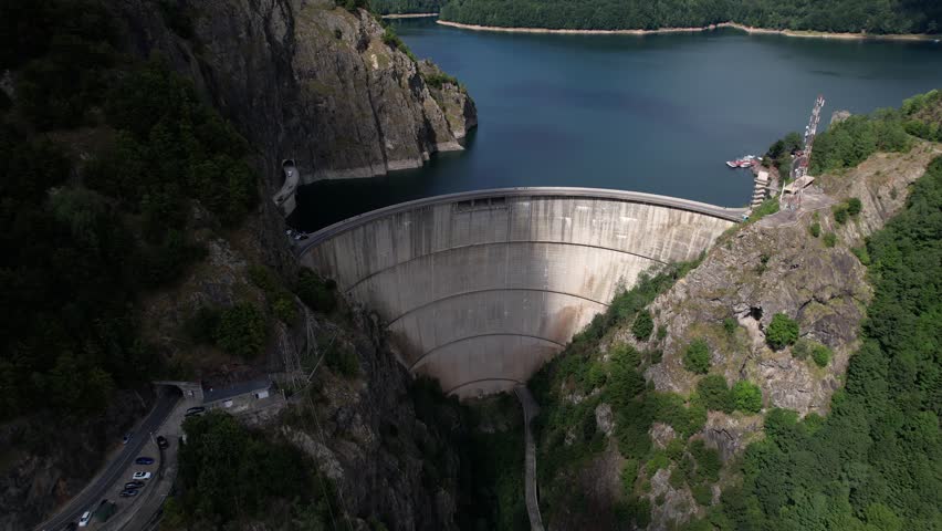 Aerial shot revealing Vidraru Dam and lake in Romania. Blue lake, dramatic curves, and green mountain backdrop in 4K.