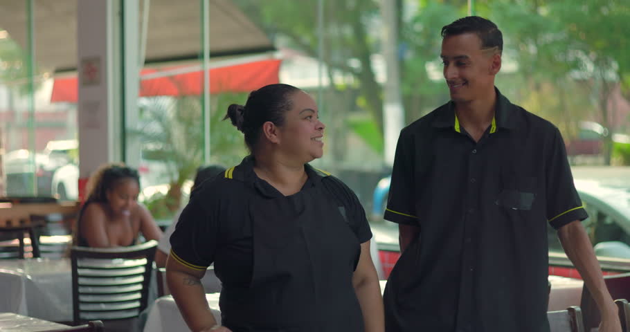 Latina waitress and young male waiter smiling together in restaurant dining area, standing side by side in friendly and welcoming pose