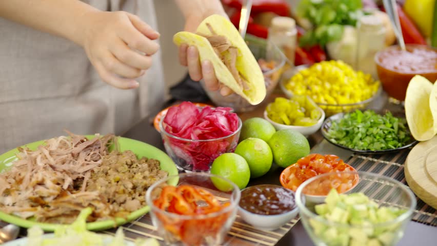 Close-up of a hand holding a plate with a fresh taco filled with avocado, herbs, and vegetables. Mexican street food served in a casual dining setting with a colorful blurred background