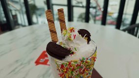 Colorful vanilla milkshake topped with whipped cream, cookies, wafer sticks, and sprinkles on a cafe table. Next to it is a glass of juice with a straw. Bright, fun, and refreshing dessert drink - Powered by Shutterstock - Get 15% off with code: PIKWIZARD15