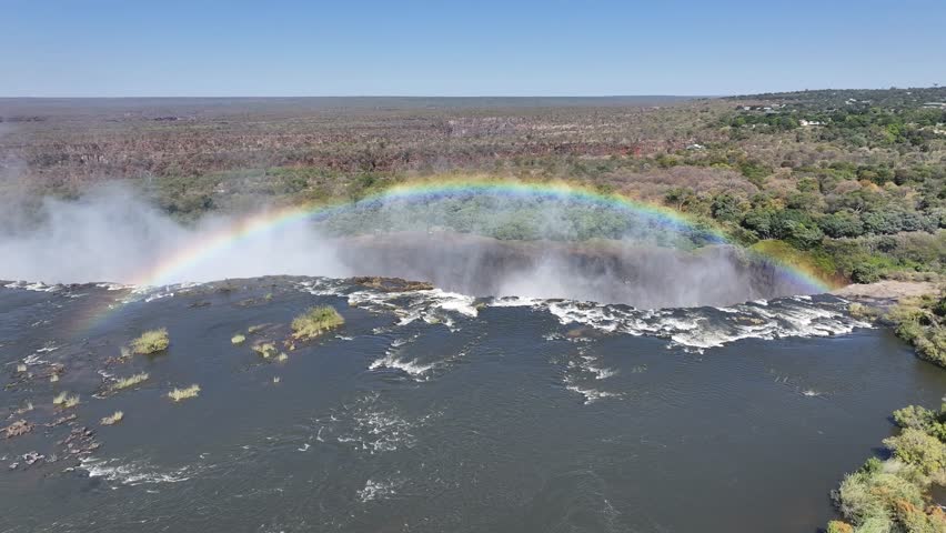 Colored Rainbow In Livingstone Northern Rhodesia Zambia. Rainbow Forming Over Waterfalls With River In Background. Nature Clouds Sky Mountain Canyon. Nature Outdoors Waterfall Powerful Flow.