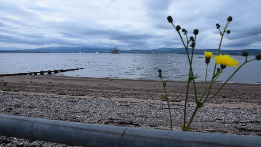 Wide shot of the Cromarty Firth in Scotland on an overcast day encompassing a beach and some hills and an oil rig in the background.