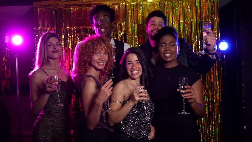 Cheerful friends are celebrating, raising glasses of champagne, posing together in a photo booth with shiny golden background
