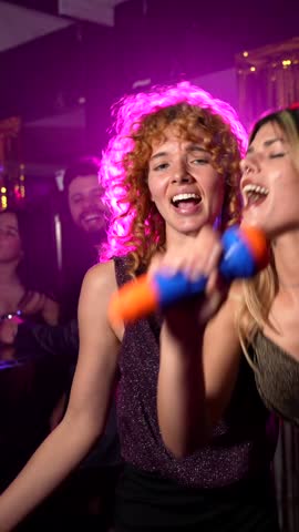 Group of young, stylish women performing energetic karaoke duet, laughing and singing together with microphones at vibrant nightclub, sharing joyful musical moment