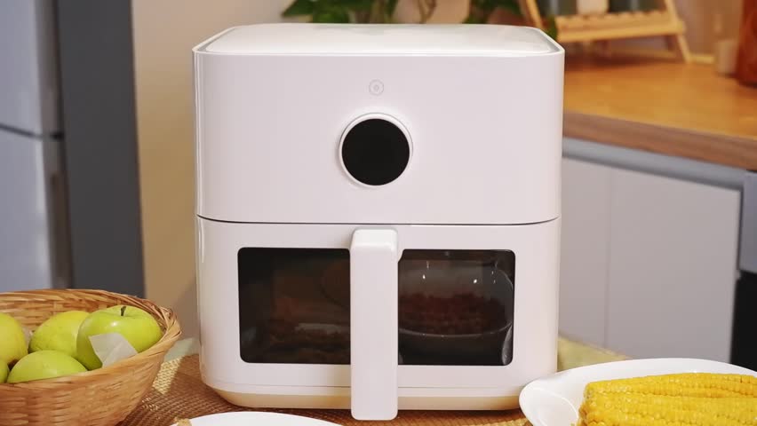Modern kitchen counter showcasing a white air fryer alongside plates of fried chicken, beans, corn, green beans, french fries, and a basket of green apples