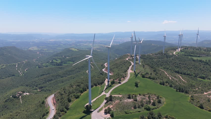 Drone shot panning slowly across a wind farm on mountain ridges, with tall wind turbines spinning under a clear blue sky and surrounded by green forest and farmland