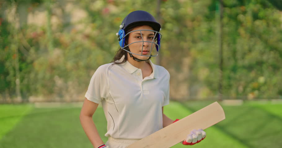 Indian gen z athlete sports woman standing at outdoor play ground wear white uniform and protective helmet looking at camera. Asian cricket player female hold bat day time at match playground