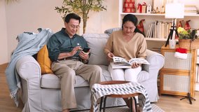 Elderly Asian couple sitting on a white sofa in cozy living room, the man using smartphone while woman flips through a book, enjoying peaceful Christmas time together at home, holiday, grandparent. - Powered by Shutterstock - Get 15% off with code: PIKWIZARD15