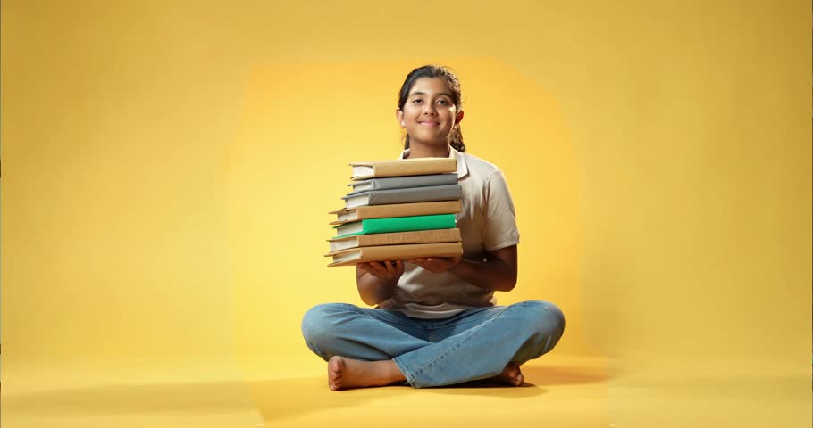 Holding Books Pile, Indian Asian Teenage Girl Sitting on Yellow Seamless Background Happily Pointing at Heap of Education Books, Promoting Learning and Study with Bright and Cheerful Expression