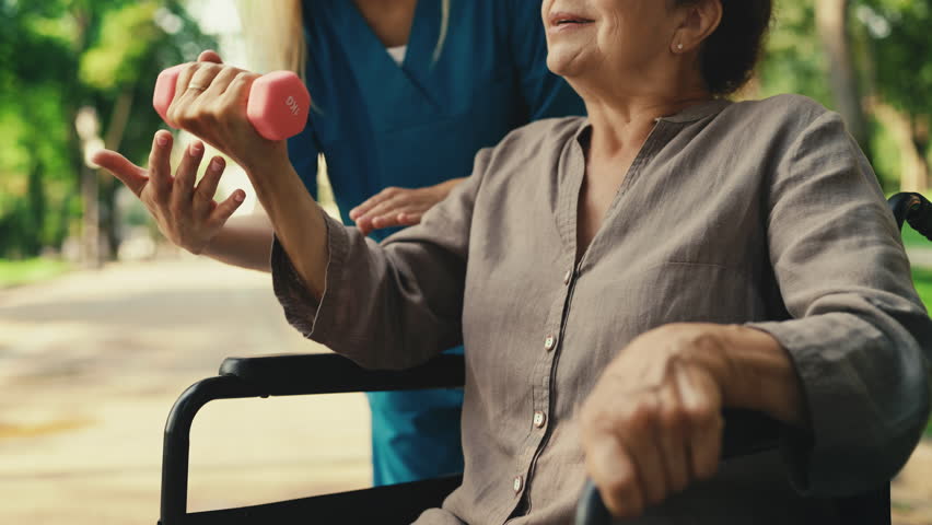 Nurse helping elderly woman in wheelchair do exercises outside, rehabilitation