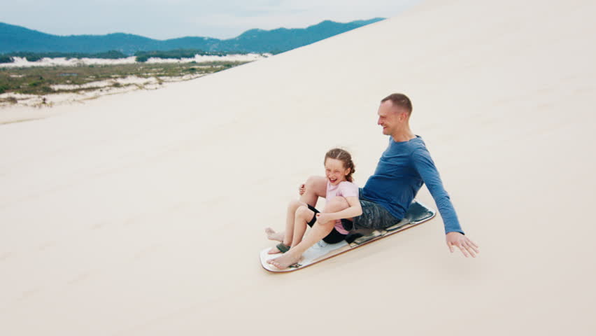 Family sandboarding. Man glides with girl down the sand dune with a lot of joy