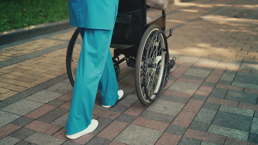 Professional nurse pushing wheelchair with senior patient, walking in city park