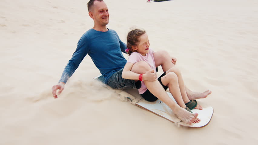 Family sandboarding. Man glides with girl down the sand dune with a lot of joy