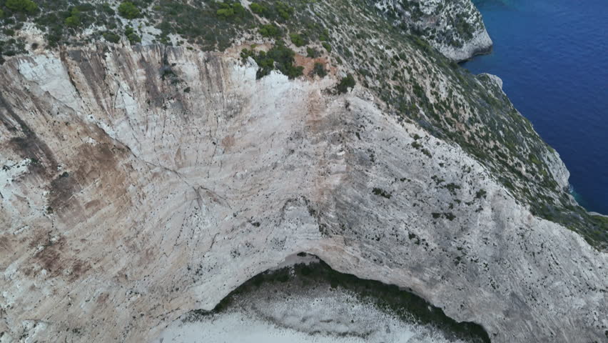 Descending aerial view over a white sandy beach framed by steep limestone cliffs, revealing two shipwrecks resting on the shore beside turquoise Mediterranean waters. The slow vertical drone movement highlights the contrast between bright sand, rugged rock formations and clear blue sea in a dramatic coastal landscape in Greece.