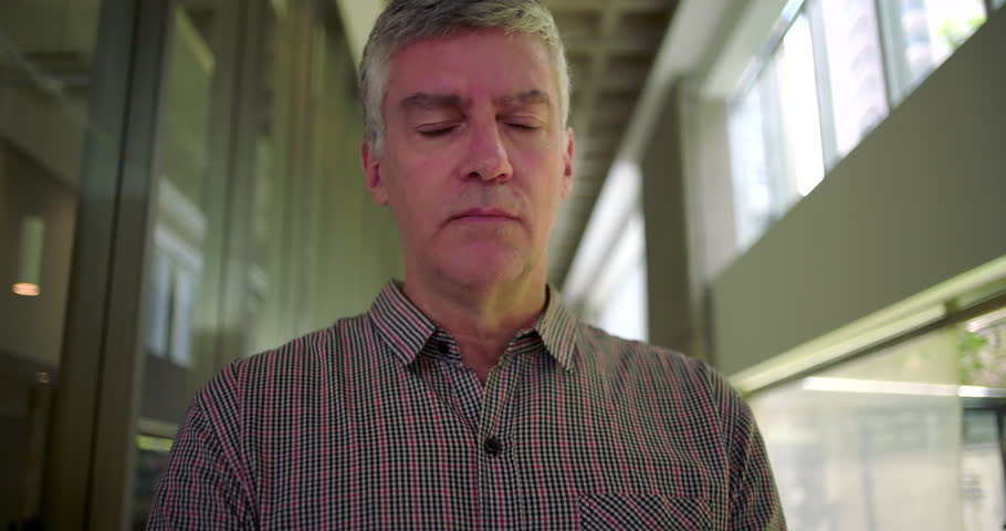 Middle aged man in office hallway looking down with tense mouth showing visible worry anxiety and mental preoccupation during moment of stress in business setting