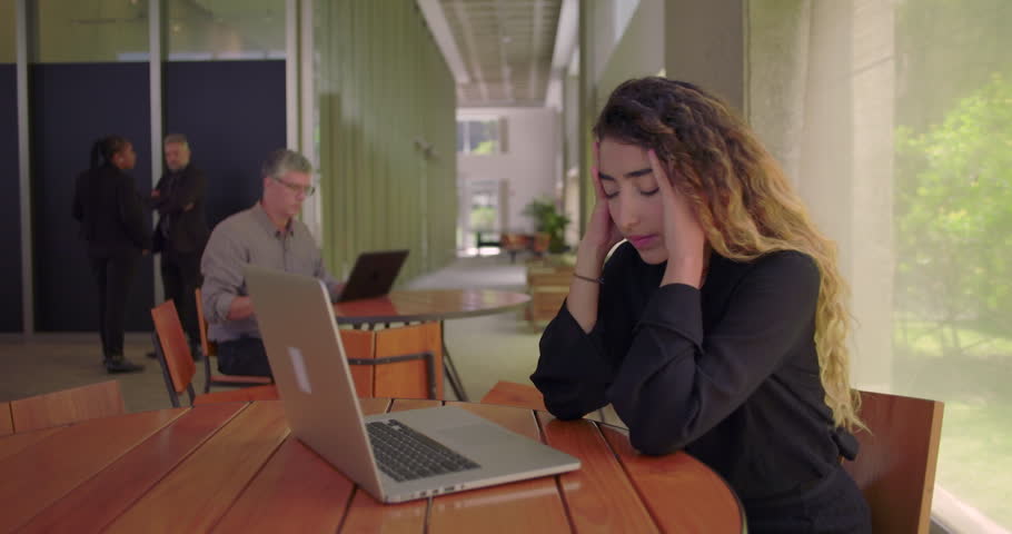 Woman covering face with hand while sitting at laptop, showing visible signs of stress and frustration in modern office, overwhelmed by workplace pressure