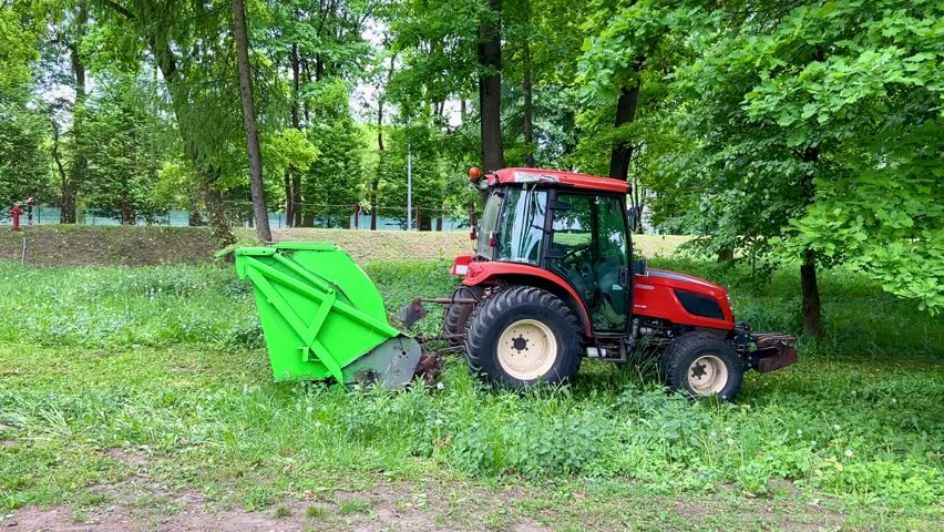 Red tractor with green mower attachment cutting grass in lush green park area during summer maintenance work.