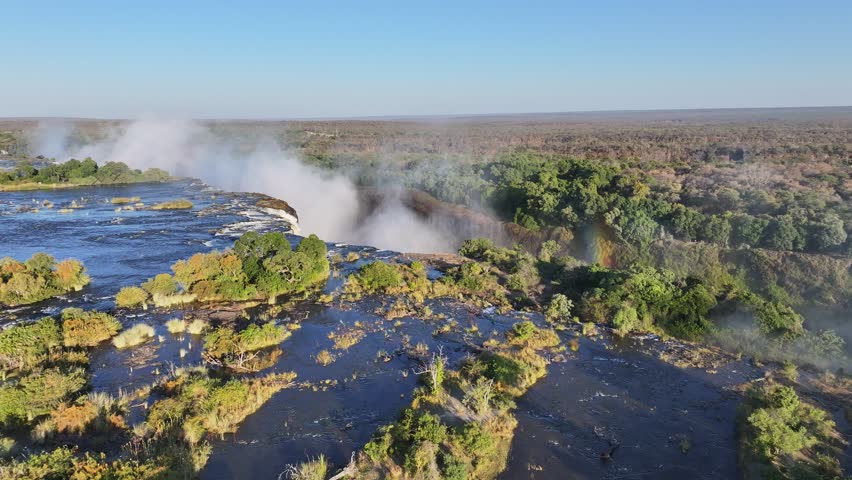 Scenic Water Fall In Victoria Falls Matabeleland North Zimbabwe. Rainbow Forming Over Waterfalls With River In Background. Landscape Sky Clouds Waterfall Tropical.