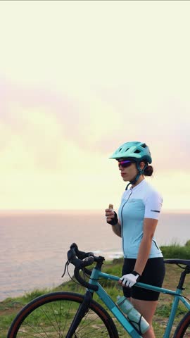 vertical Latin cyclist woman wearing helmet and technical clothing eating an energy bar standing next to her gravel bicycle in front of the sea at sunrise