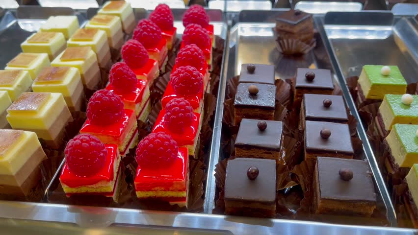 Assortment of colorful mini pastries with chocolate, raspberry fruit, and mousse layers on display in a bakery showcase, neatly arranged in rows